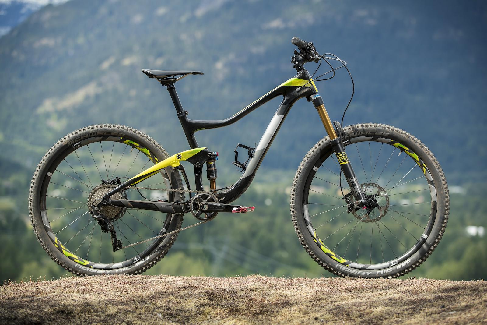 Giant Giant Trance Advanced 27.5 2: Image of a modern mountain bike with a sleek black and yellow frame, positioned on a rocky surface with a blurred mountainous landscape in the background. The bike features thick tires suitable for off-road terrain and a front suspension fork, emphasizing its design for rugged trails and adventure riding.