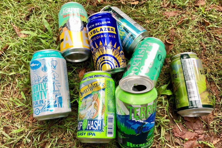A collection of colorful beer cans scattered on green grass, including brands like Lagunitas, New Belgium, Dogfish Head, SweetWater, and Terrapin. The cans feature various designs and labels indicating different styles of beer, such as IPAs and session ales. Some cans are partially crushed, adding to the casual outdoor setting.