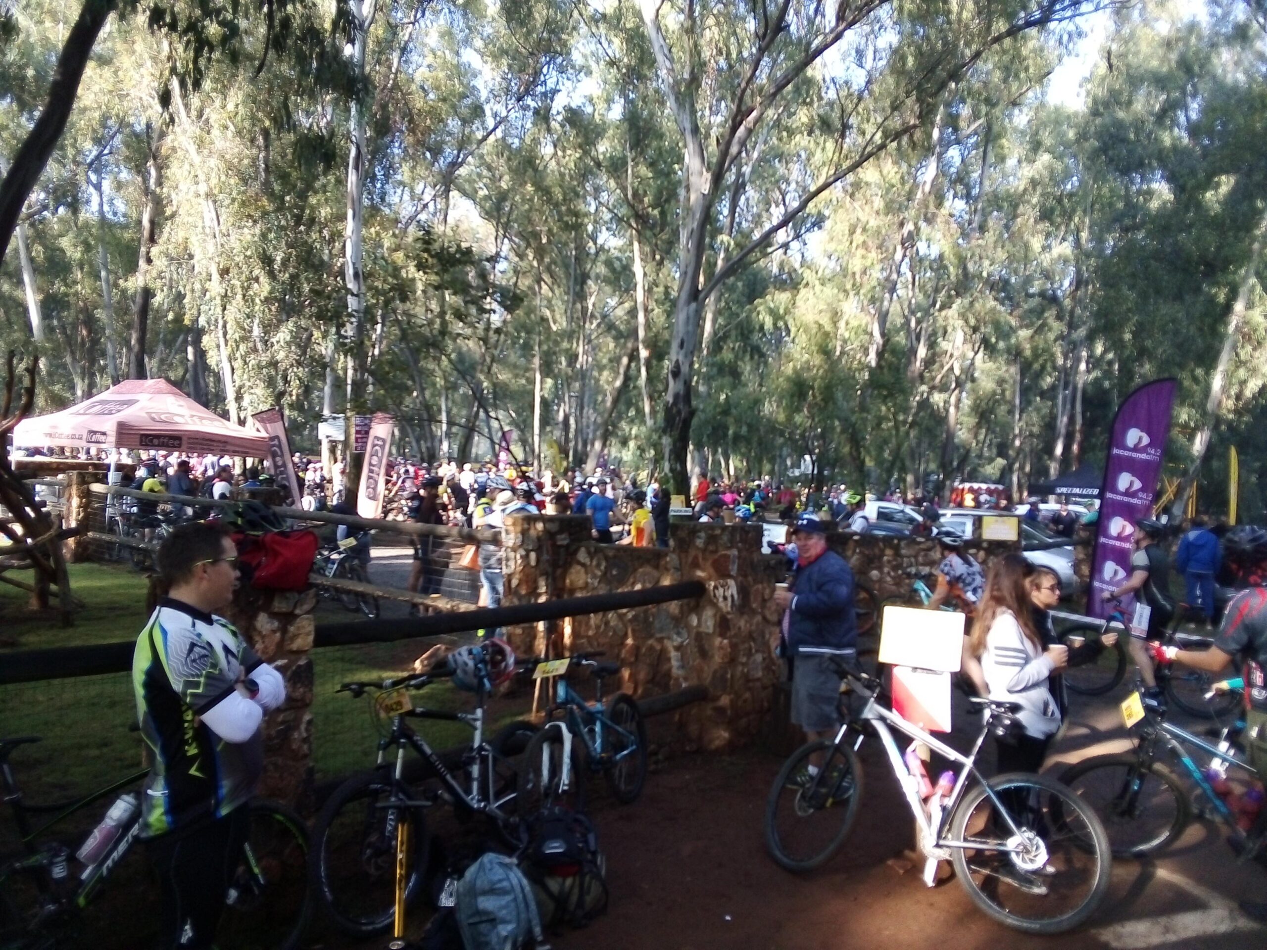 GT Helion Comp: A lively outdoor scene depicting a mountain biking event. Groups of participants and spectators gather among tall trees, with a tent in the background offering refreshments. Bicycles are parked nearby as people socialize, showcasing a vibrant atmosphere of community and sportsmanship.