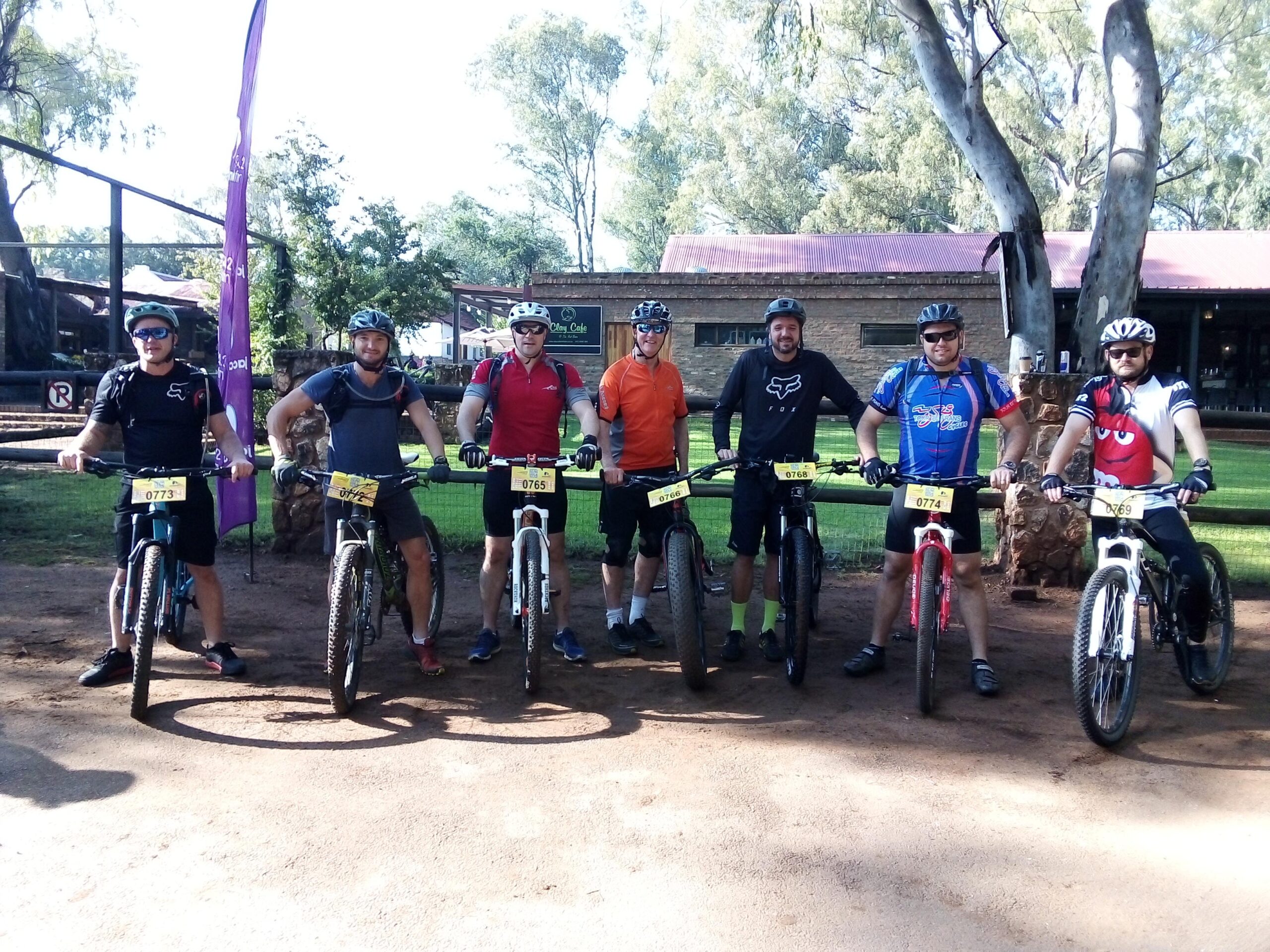 GT Helion Comp: Seven mountain bikers stand side by side with their bikes in a park-like setting. They are wearing helmets and various cycling jerseys, and they appear ready for a biking event. In the background, there are trees and a building with a red roof. The scene captures a sunny day and an atmosphere of camaraderie and anticipation.