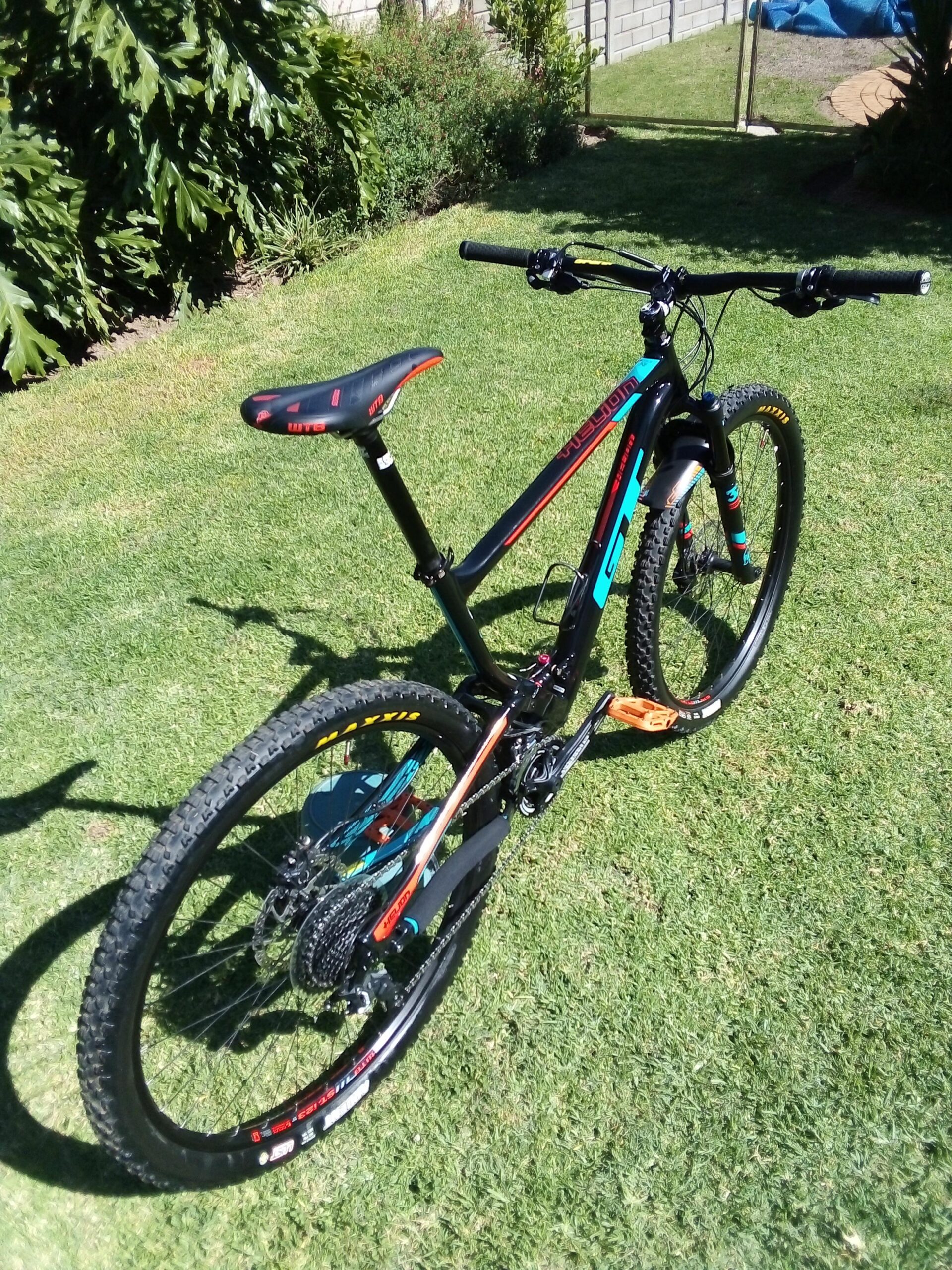 GT Helion Comp: A black mountain bike with colorful accents is positioned on green grass. The bike features a sleek design with a prominent seat and wide handlebars. The tires have a rugged tread pattern suitable for off-road riding, and the background includes lush plants and a paved pathway.