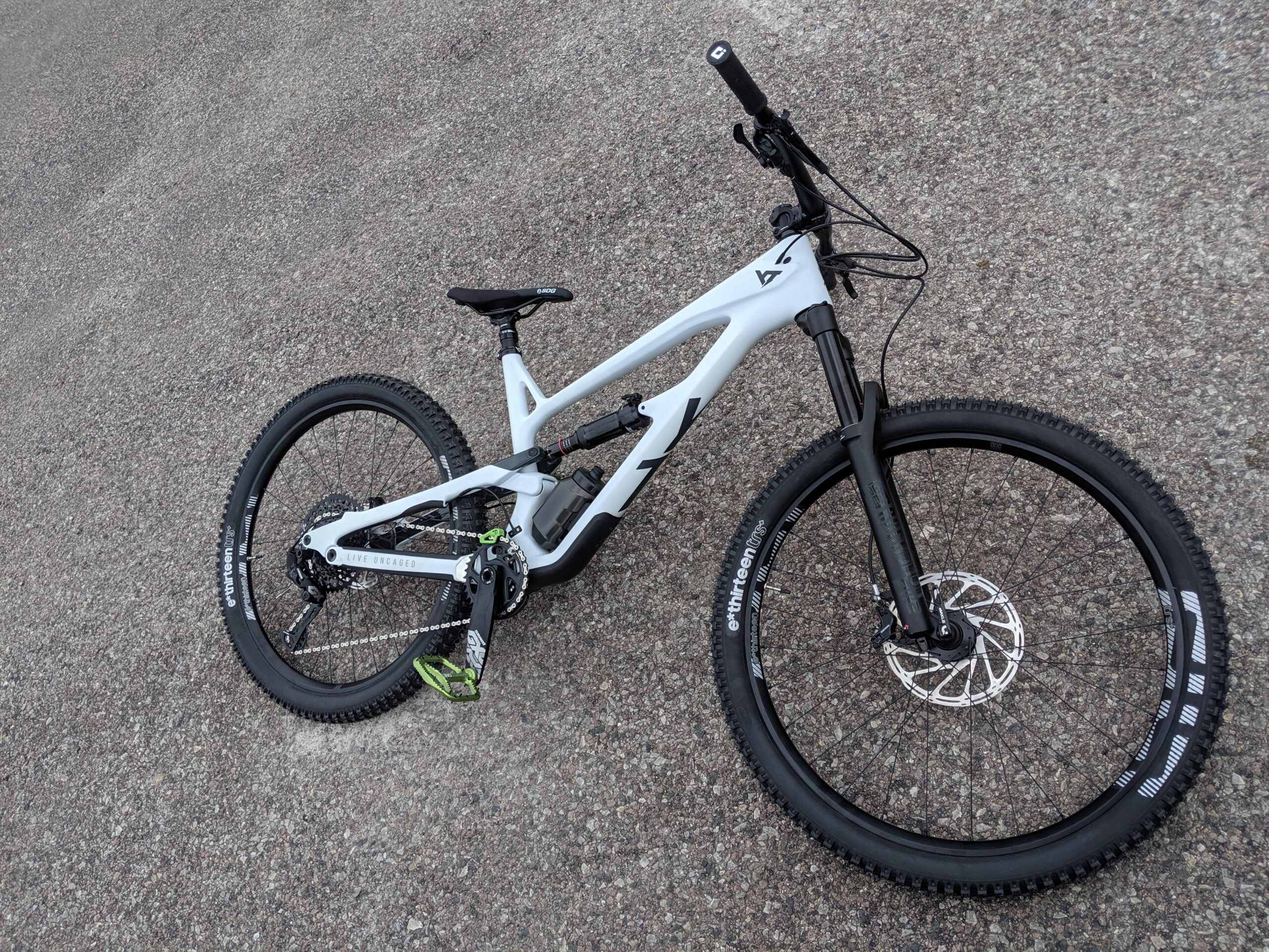 YT Industries YT Jeffsy CF: Alt text: A white mountain bike is positioned on a gravel surface. The bike features a black fork, thick tires with tread patterns, and components including a gear shifter and pedals. The frame has bold markings and a sleek design.
