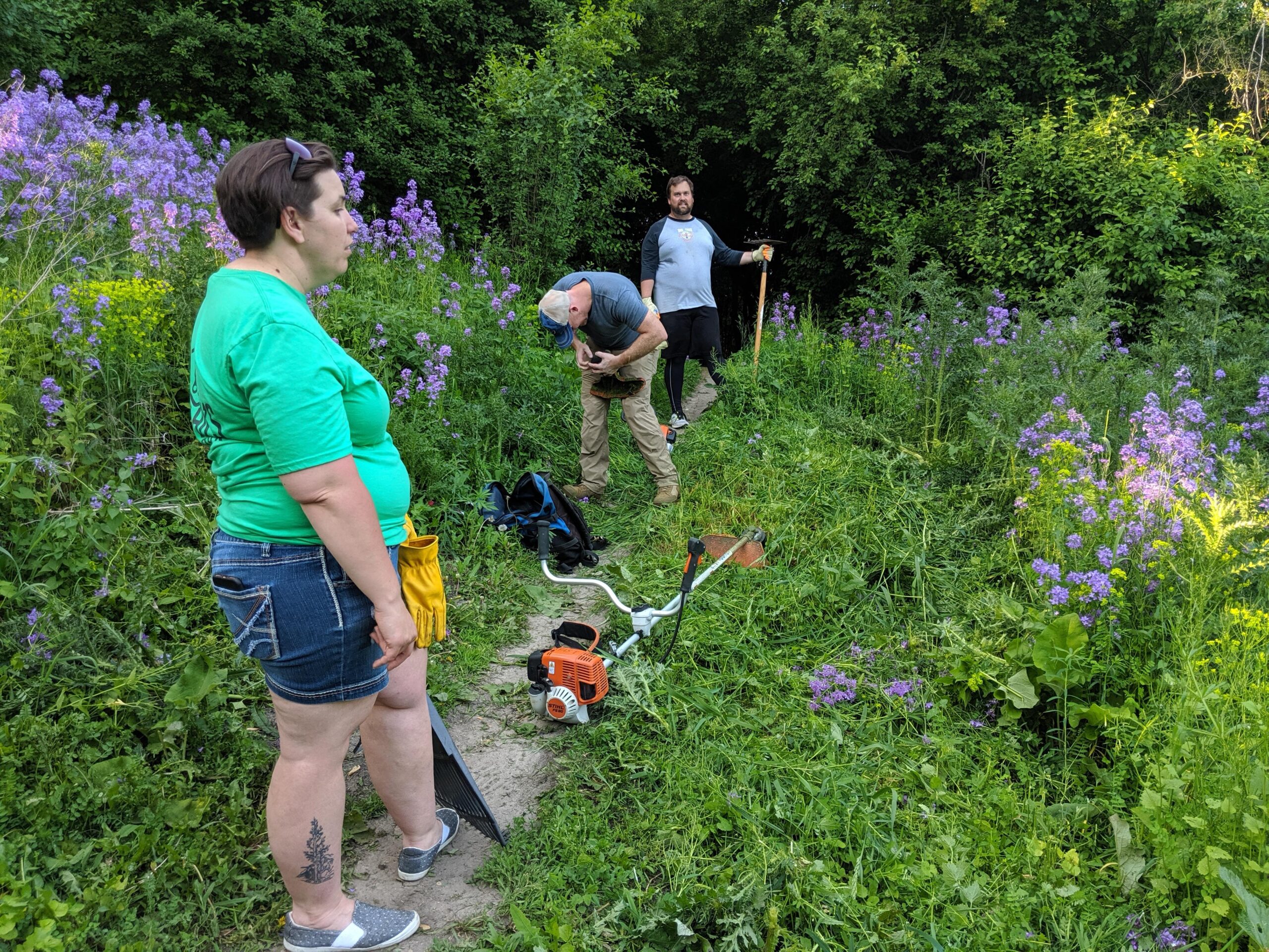 A group of three individuals working in a lush outdoor setting filled with purple flowers. One person stands in profile wearing a green shirt and denim shorts, while another person, focused on their task, is bent over inspecting equipment. A third person stands nearby with a gardening tool. The scene is vibrant with greenery and flower blooms.