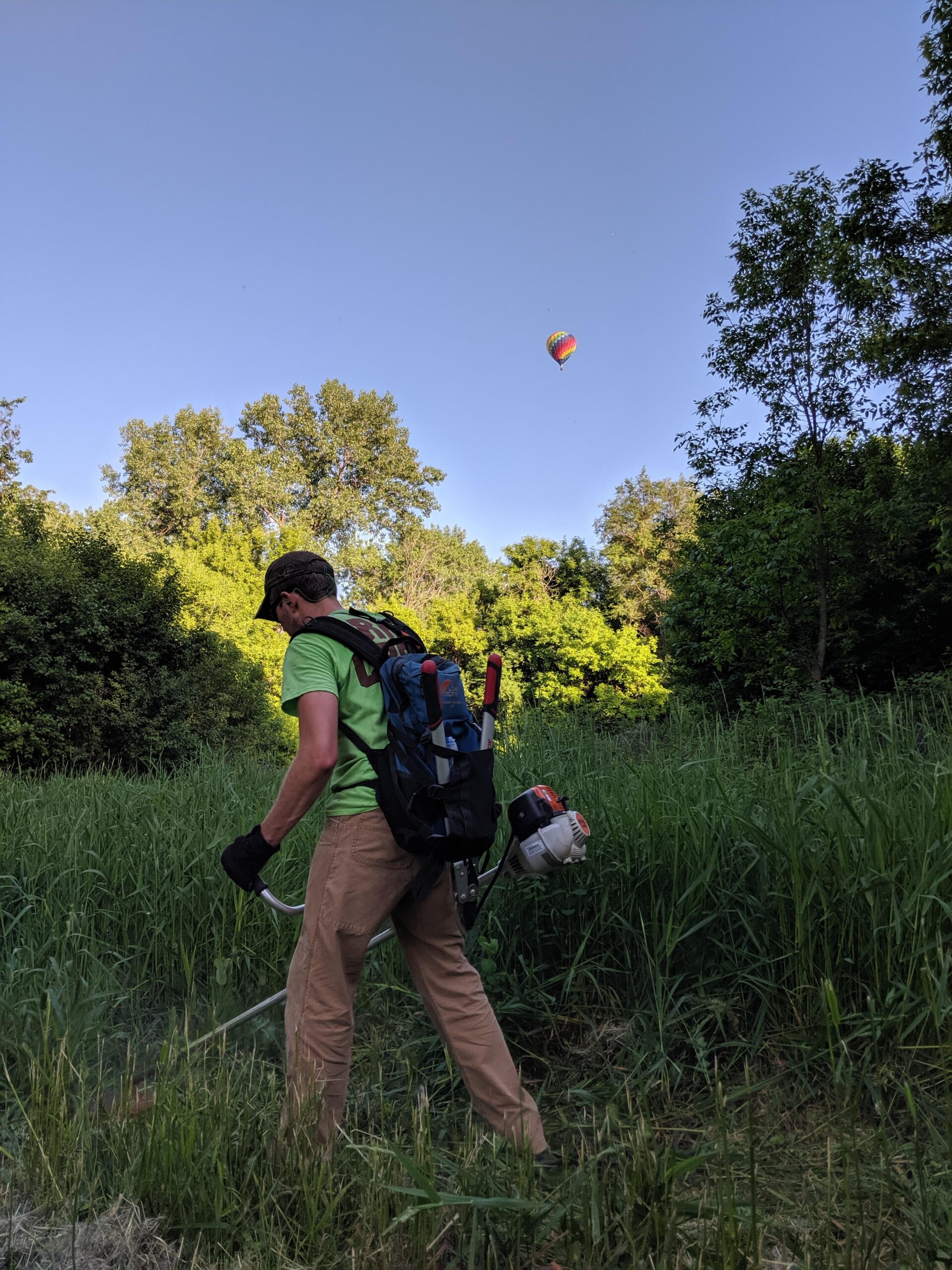 A person wearing a green shirt and tan pants is using a string trimmer in a grassy area. They have a backpack mounted with tools and equipment, and a colorful hot air balloon can be seen flying in the blue sky above a backdrop of trees.