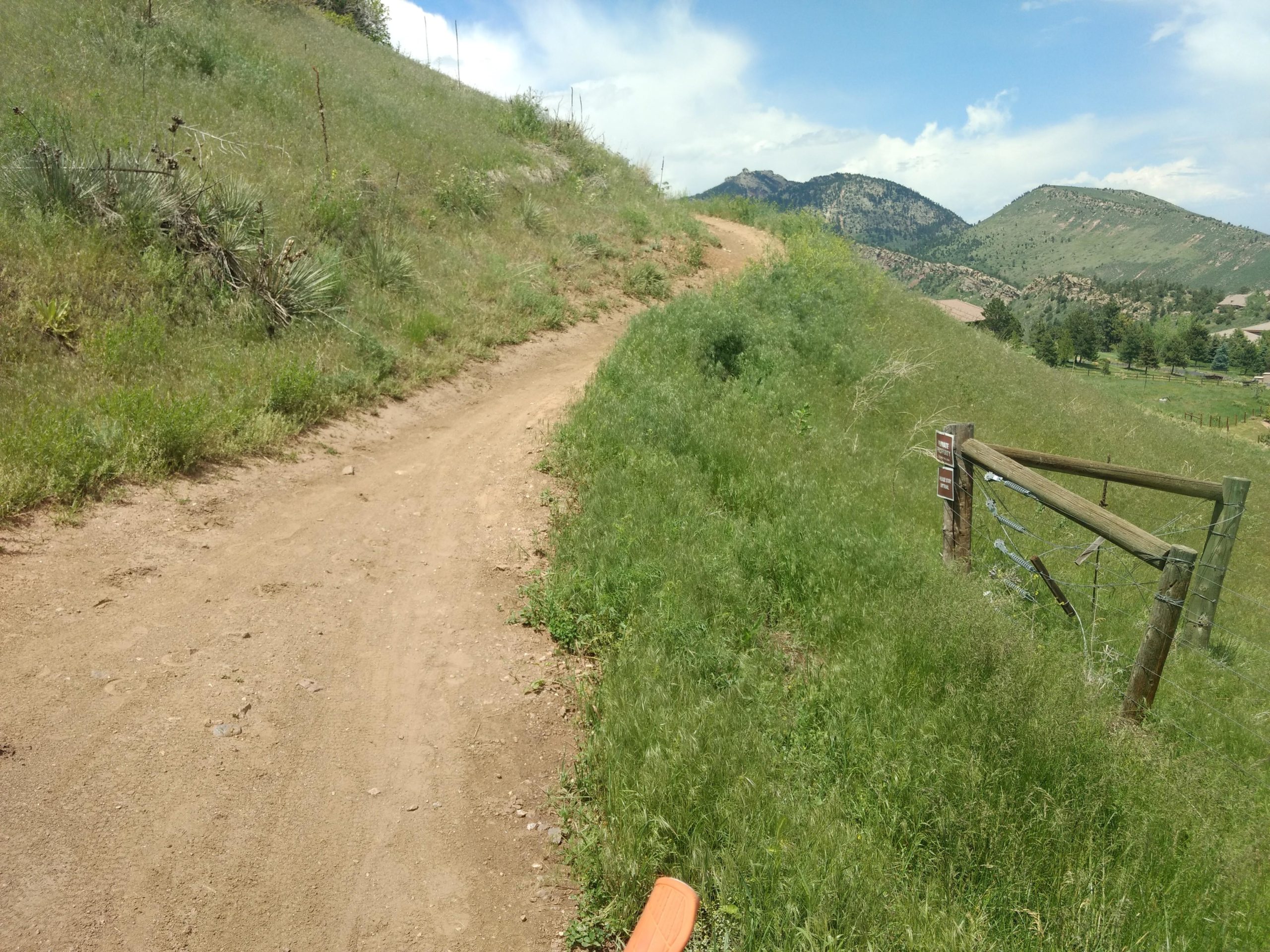 A dirt path curves through green, grassy hills under a partly cloudy sky, with mountains visible in the background. A wooden fence with a sign is to the right, and the landscape is dotted with shrubs and wild plants. White Ranch mountain bike trail.