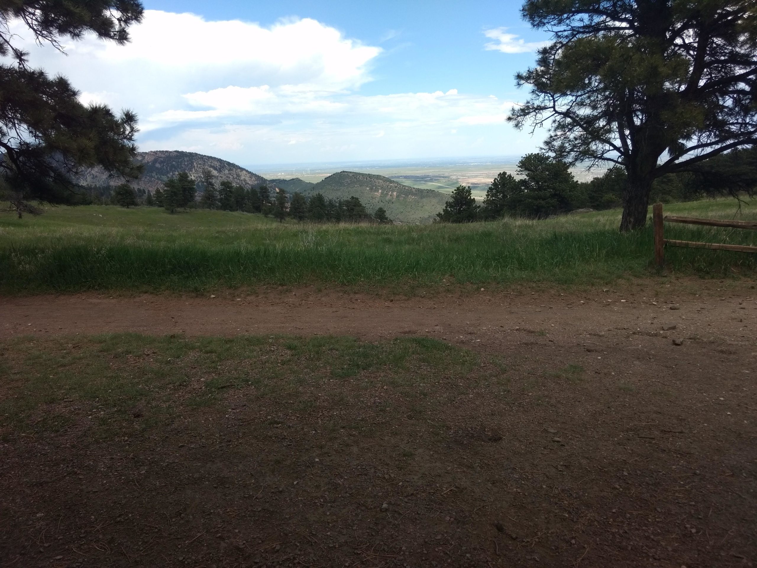 A scenic view of rolling hills and a grassy field under a partly cloudy sky. In the foreground, a dirt path leads into the lush greenery, while trees frame the edges. The background features distant mountains that gently rise against the horizon, with a valley visible in the distance. White Ranch mountain bike trail.
