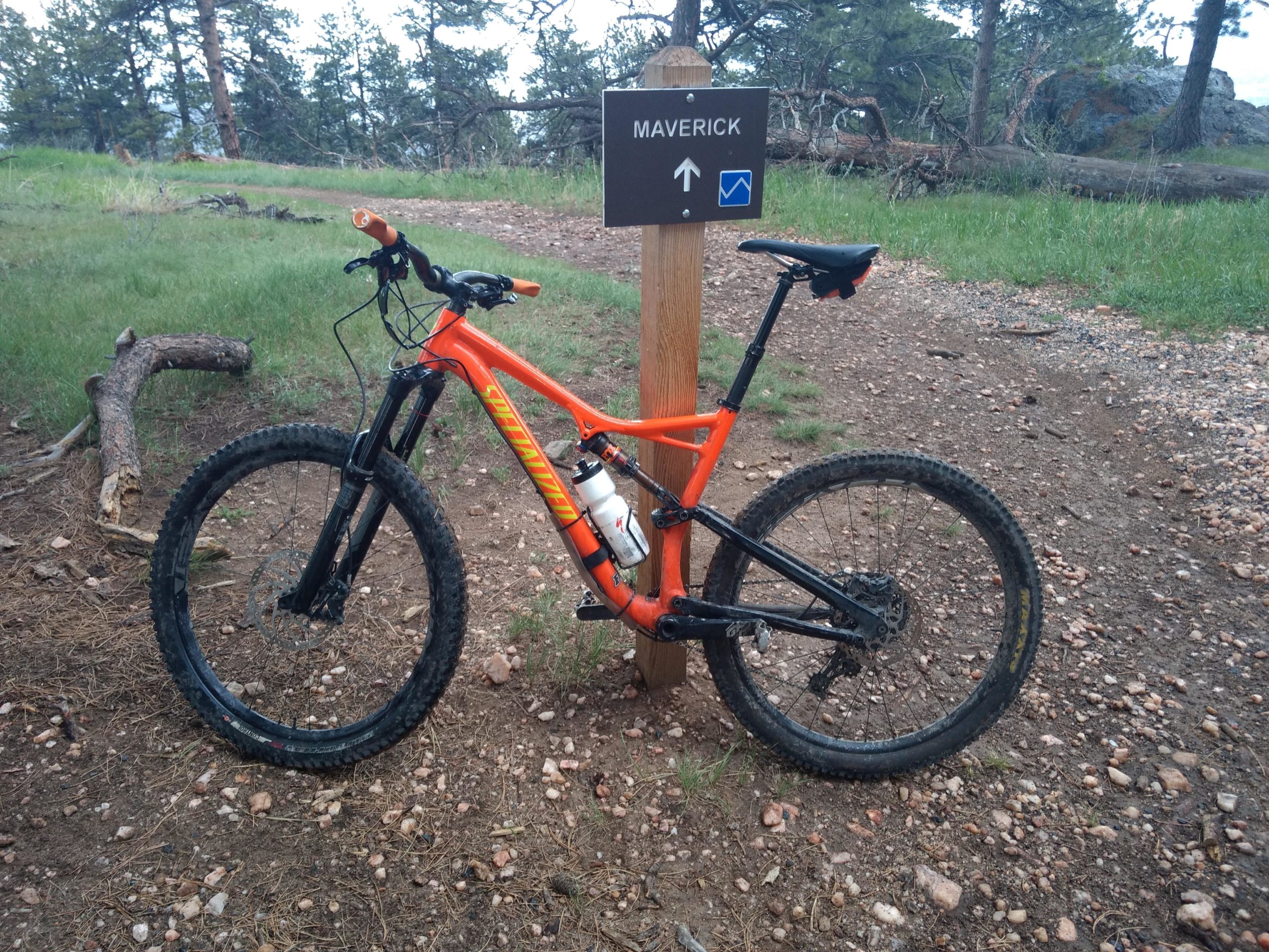 A bright orange mountain bike rests next to a wooden sign indicating the trail name "Maverick," with an arrow pointing in the direction of the trail. The scene is set in a wooded area with rocky terrain and a mix of grass and dirt. Trees are visible in the background, suggesting a natural outdoor environment. White Ranch mountain bike trail.