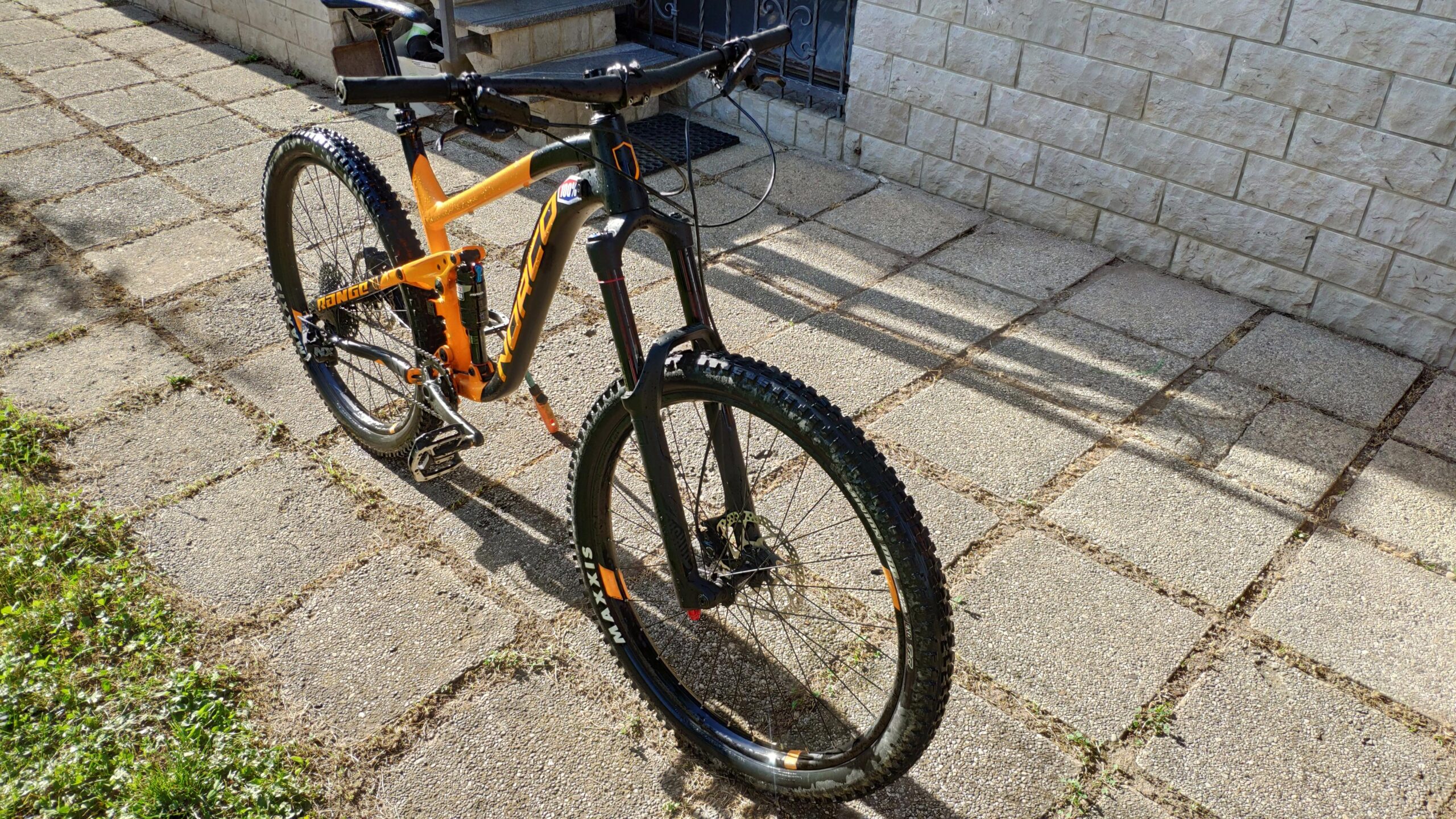 Norco Norco Range A7.3: A vibrant orange mountain bike parked on a stone pavement. The bike features a front suspension fork and rugged tires suitable for off-road cycling. Sunlight casts shadows on the ground, highlighting the bike's sleek design and sturdy frame. In the background, a wall made of light-colored stone is partially visible.
