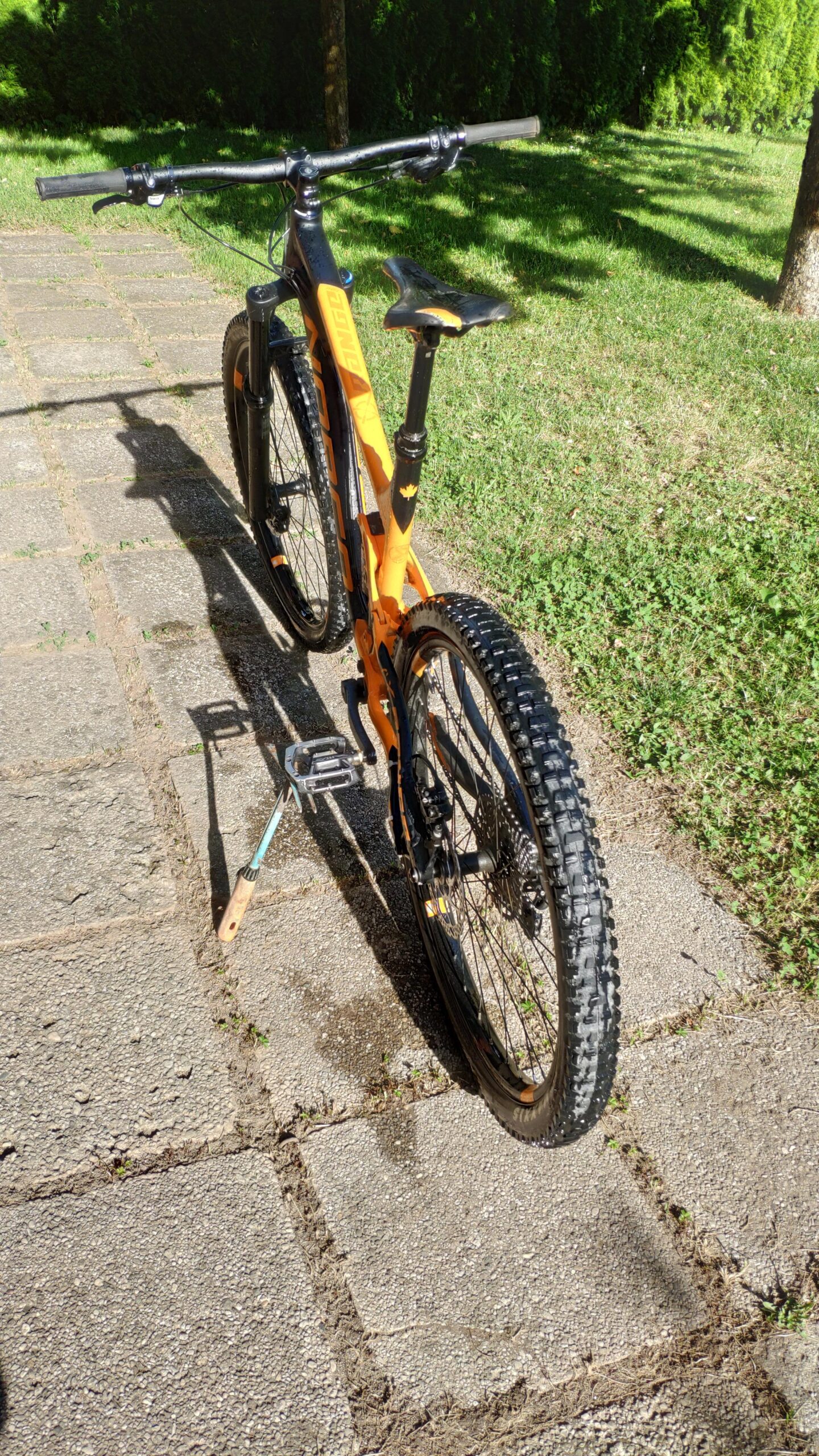 Norco Norco Range A7.3: A mountain bike with an orange frame parked on a stone path. The bike features thick, knobby tires and a black seat. Lush greenery is visible in the background, providing a natural setting.