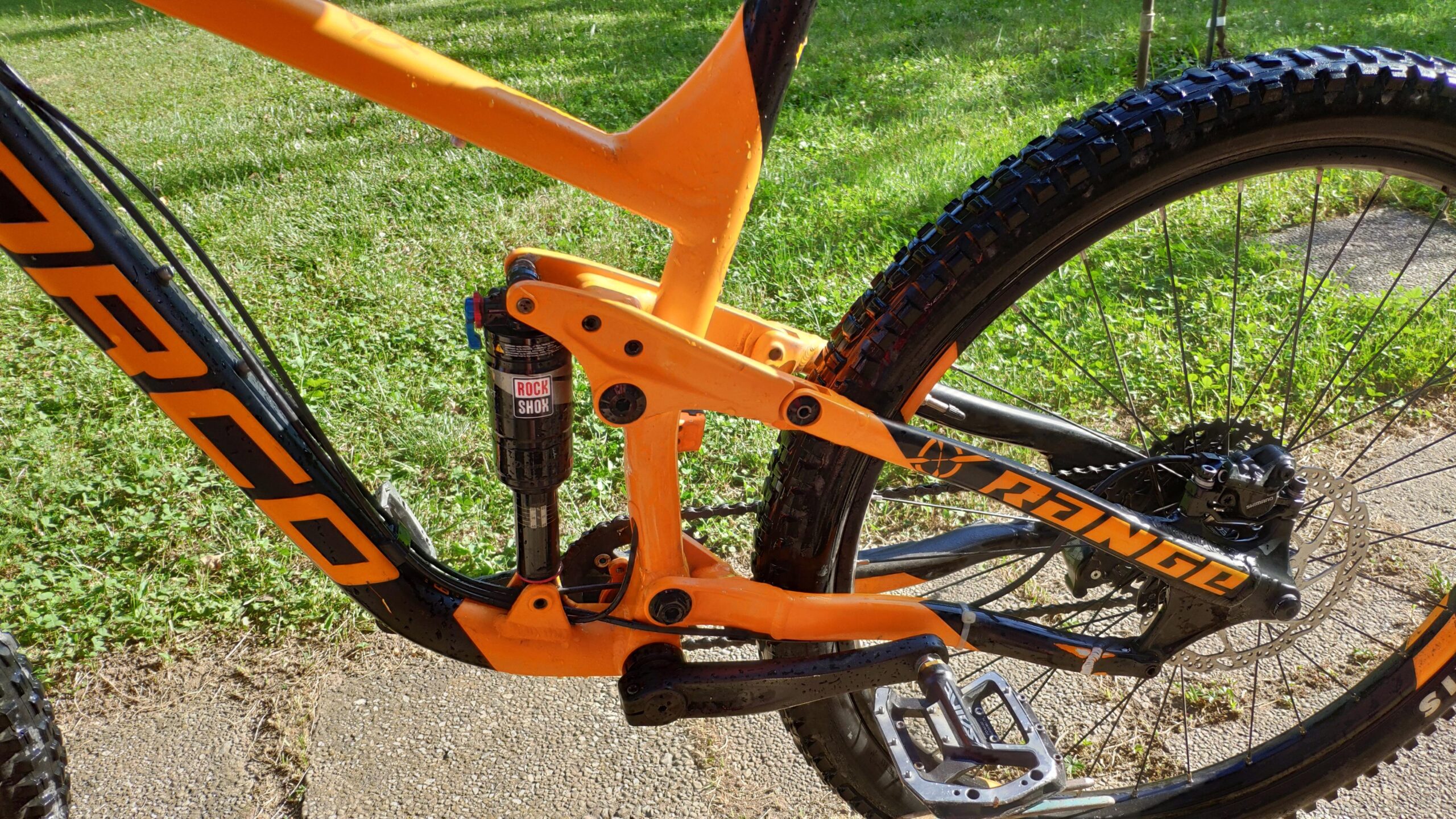 Norco Norco Range A7.3: Alt text: Close-up view of a mountain bike frame with an orange and black design, showcasing the suspension components and drivetrain, set against a grassy background.