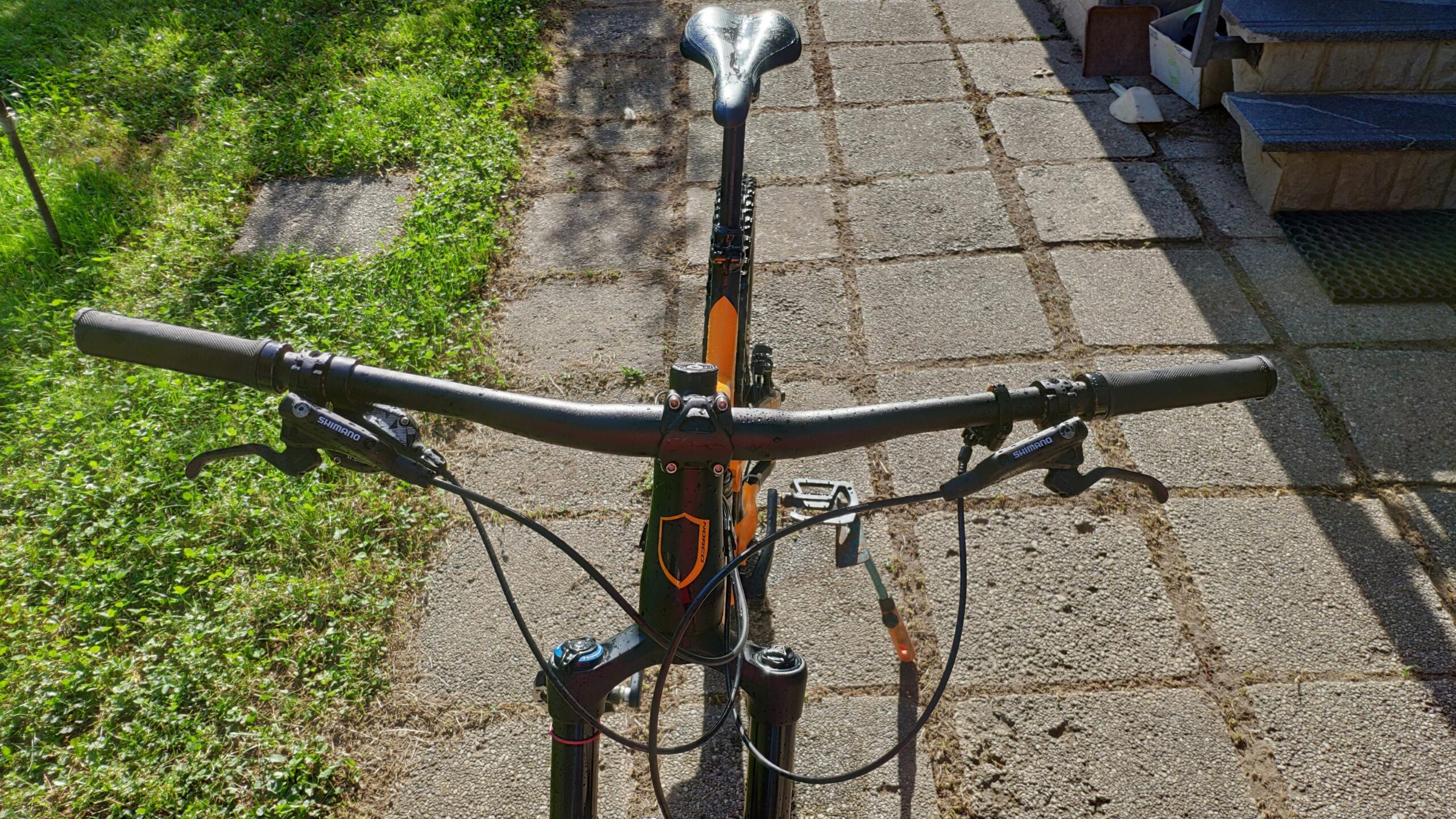 Norco Norco Range A7.3: A top-down view of a mountain bike's handlebars and front fork on a paved path, surrounded by green grass. The bike features Shimano brake levers and textured grips. Sunlight casts shadows, highlighting the bike's components and the surrounding stones.