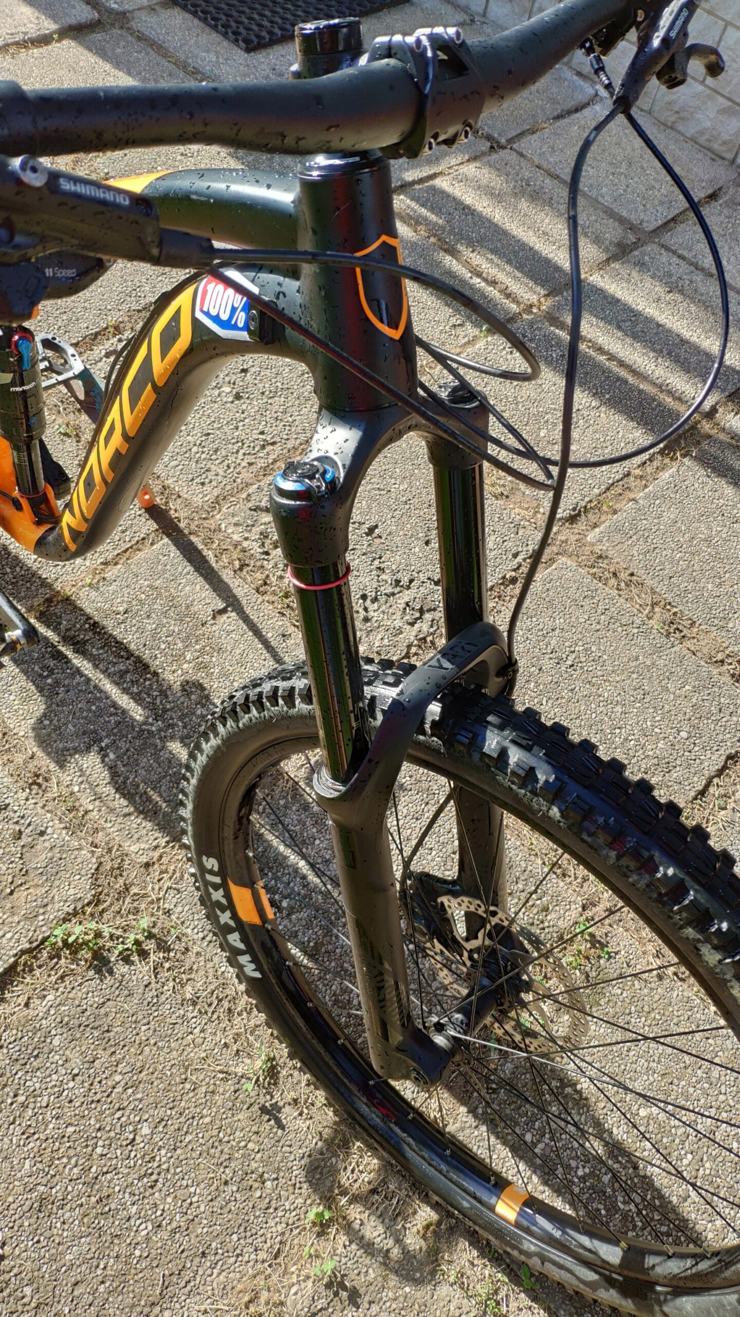 Norco Norco Range A7.3: Close-up view of a black and orange mountain bike featuring a sturdy front fork, with water droplets on the frame. The handlebars are visible along with a Shimano gear system. The tire has a textured tread suitable for off-road use, and the bike is positioned on a gravel surface with some grass peeking through.