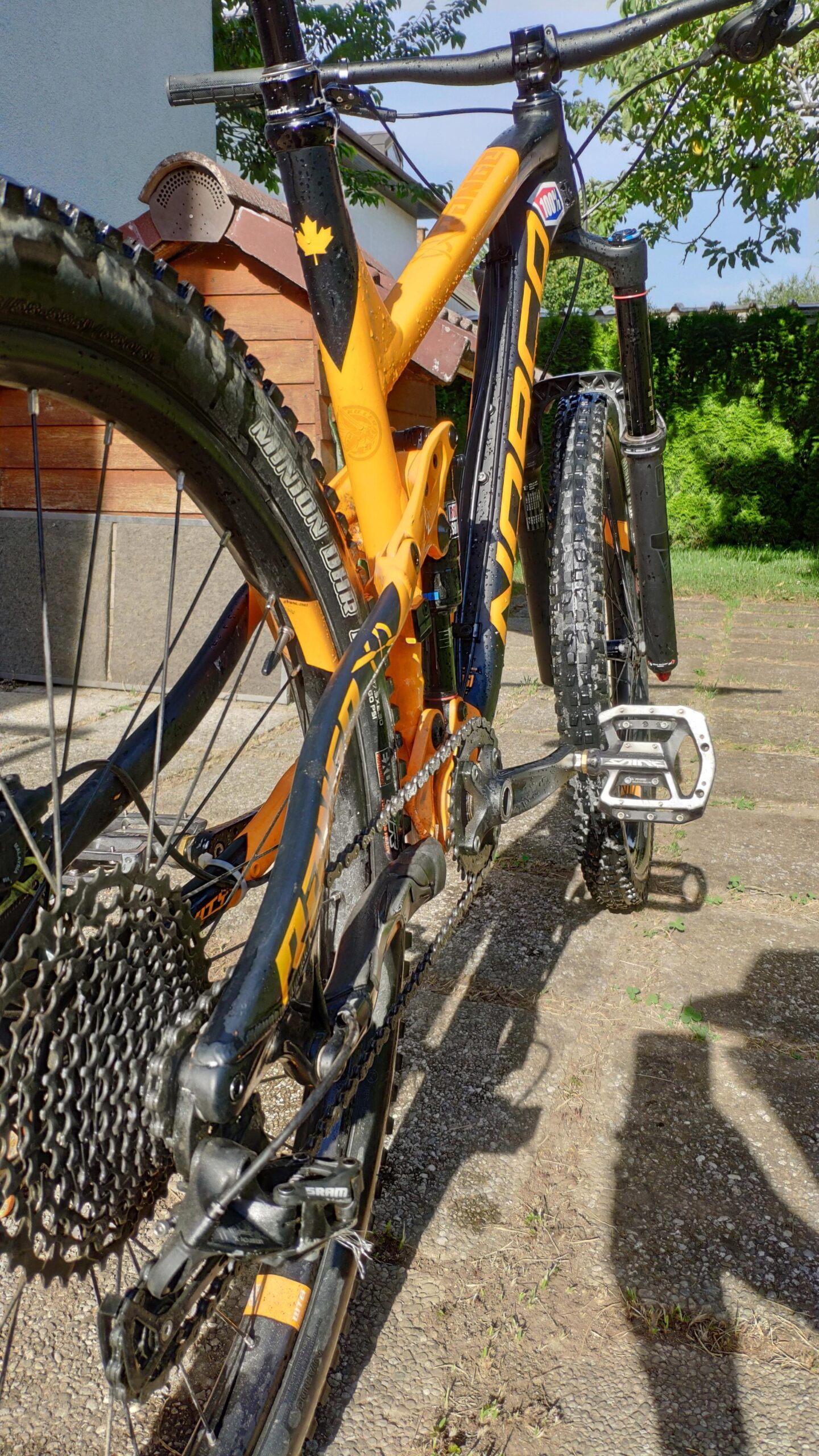 Norco Norco Range A7.3: A close-up view of a mountain bike with orange and black detailing, showcasing the rear wheel, gear system, and pedal. The bike is parked on a stone surface with greenery in the background and a wooden structure partially visible. Water droplets are present on the bike, indicating it may have just been washed or used in wet conditions.