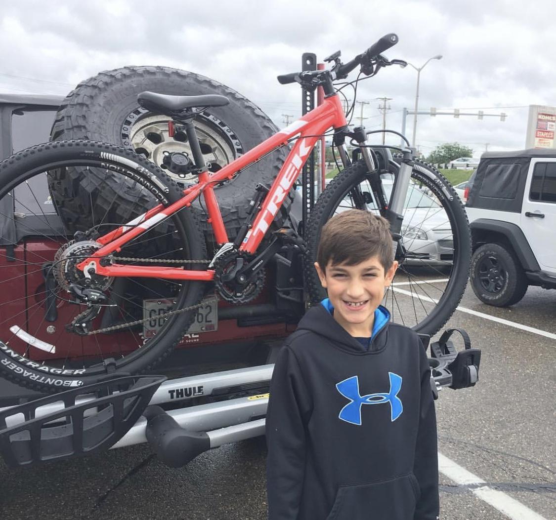 Trek Marlin 5: A young boy with brown hair smiles in front of a red vehicle, featuring a mountain bike mounted on a rack. The bike has visible branding and is positioned alongside a large spare tire. The background shows an overcast sky and a parking lot with other vehicles.