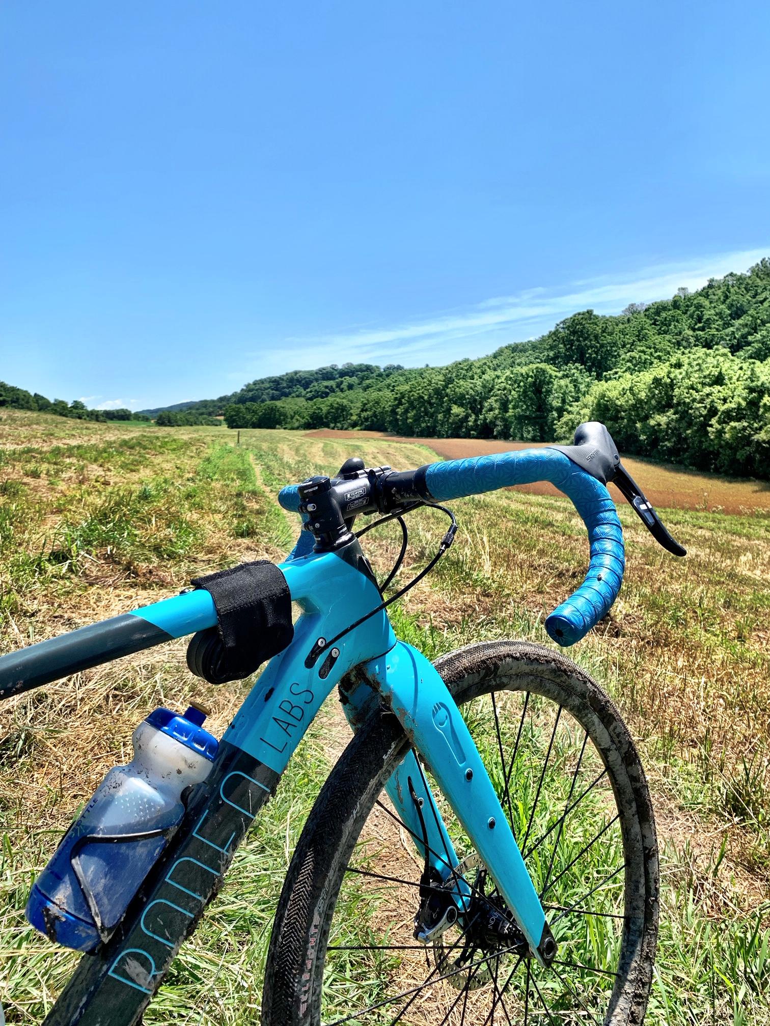 A close-up view of a turquoise bicycle parked on a grassy field, with a clear blue sky in the background. The bike features a water bottle mounted on the frame and is partially covered in mud, suggesting recent off-road use. Lush green hills can be seen in the distance. Blue Marsh mountain bike trail.