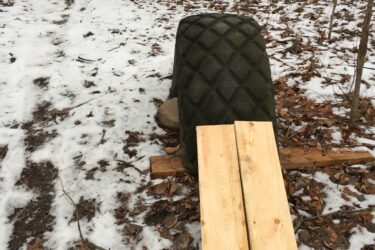 Two large rubber tires are positioned on a snowy path in a wooded area, each supported by wooden planks that extend across the ground. Fallen leaves cover the ground, contrasting with the snow. The scene captures a serene yet rugged natural environment. Swance Drain mountain bike trail.