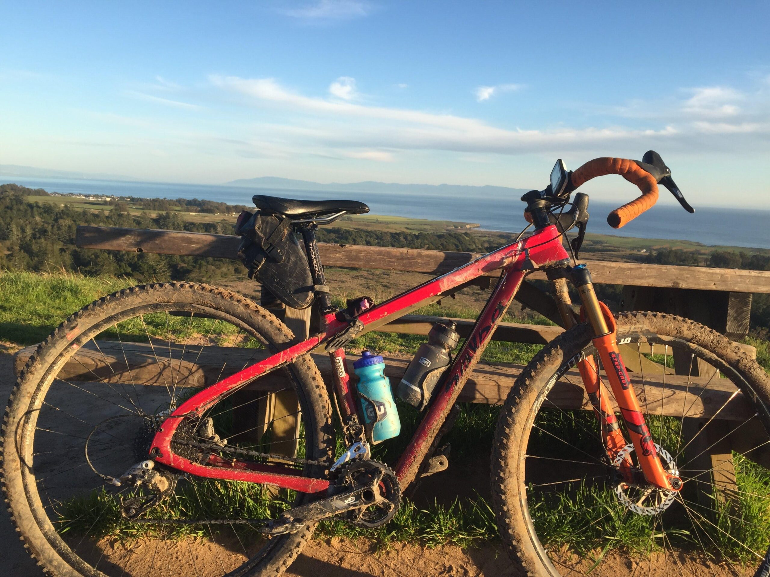 Salsa Cutthroat: A mountain bike with a red frame and orange handlebars is parked beside a wooden railing, overlooking a scenic view of a landscape with hills and a body of water in the distance. The bike features muddy tires and a water bottle attached, set against a clear blue sky.