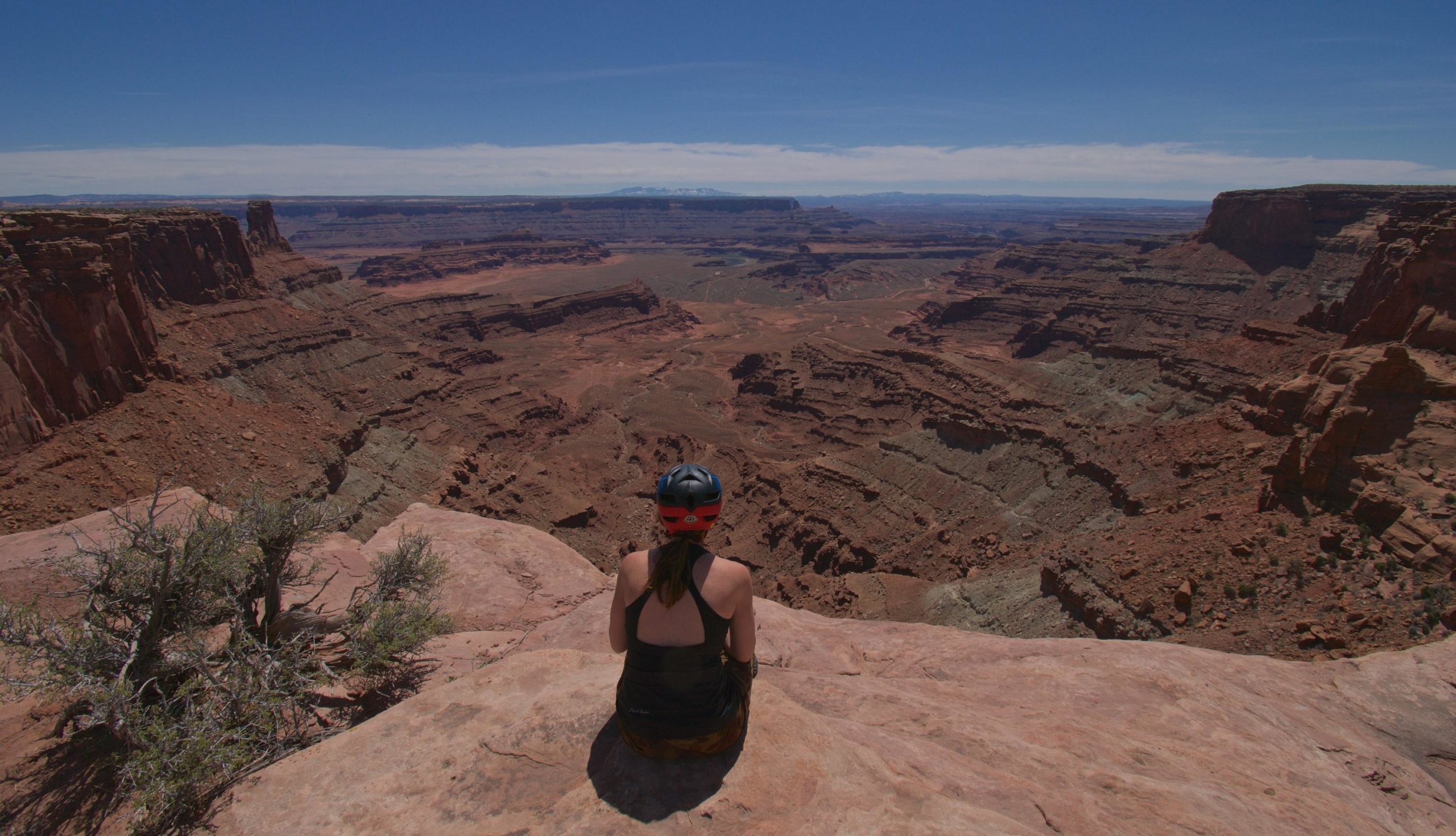 A person sitting on the edge of a rocky cliff, overlooking a vast desert canyon landscape under a clear blue sky. The scene showcases layers of red and brown rock formations, with a hint of greenery in the foreground. Dead Horse Point State Park mountain bike trail.