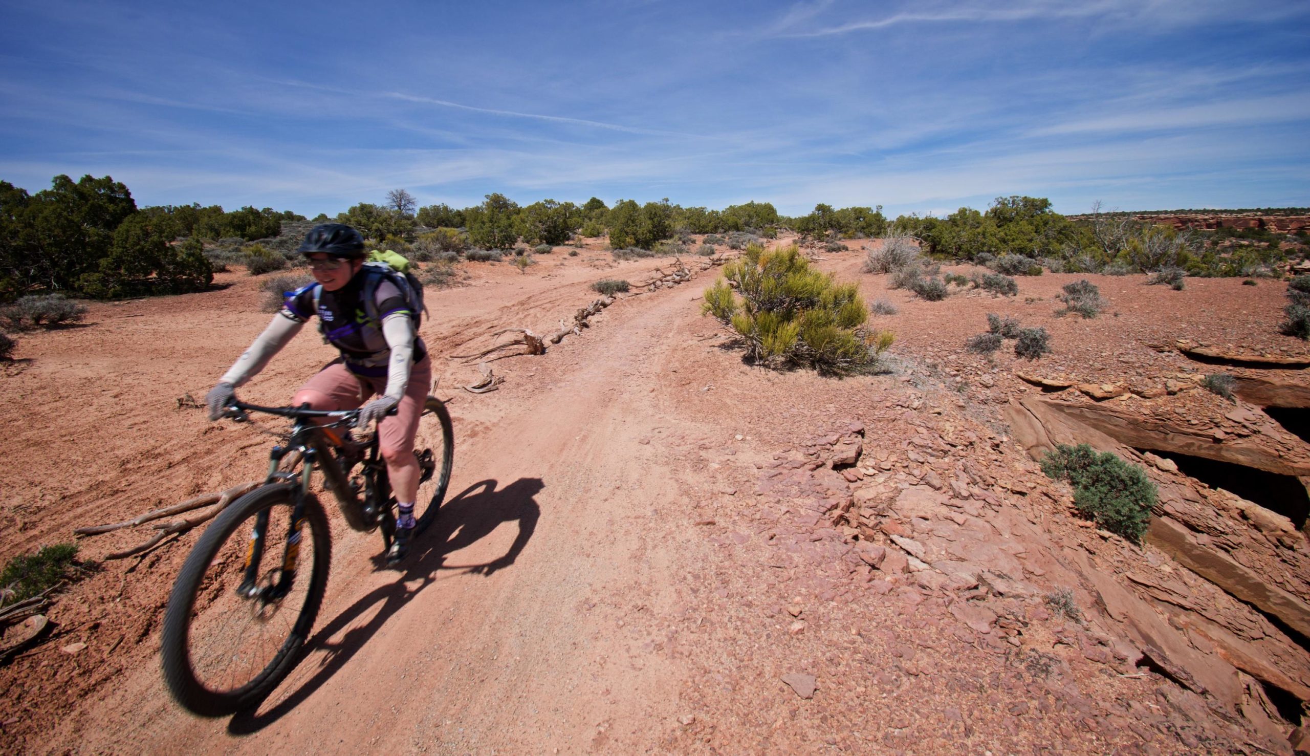 A mountain biker rides along a dirt trail in a desert landscape, surrounded by vegetation and rocky terrain under a clear blue sky. The cyclist wears a helmet and a backpack, appearing to navigate a winding path. Dead Horse Point State Park mountain bike trail.