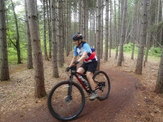 A cyclist riding a mountain bike along a narrow dirt trail in a forest, surrounded by tall pine trees. The rider is wearing a blue and red jersey, shorts, and a helmet, with a blurred motion effect indicating speed. Cuyuna Lakes mountain bike trail.