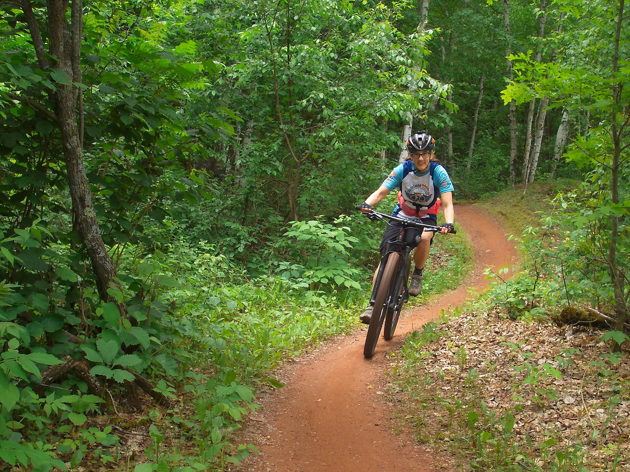 A person riding a mountain bike on a winding dirt trail surrounded by lush green vegetation and trees. The cyclist is wearing a helmet and a colorful shirt, enjoying outdoor biking in a natural setting. Sandhog Mountain mountain bike trail.