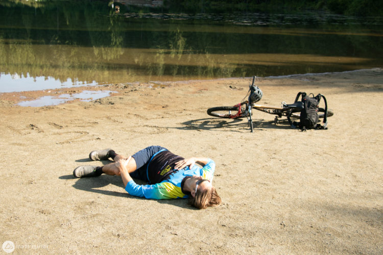 A person lying on the sandy shore near a calm body of water, wearing a blue and yellow cycling jersey and shorts. A mountain bike and backpack are situated nearby, with trees reflected in the water in the background.