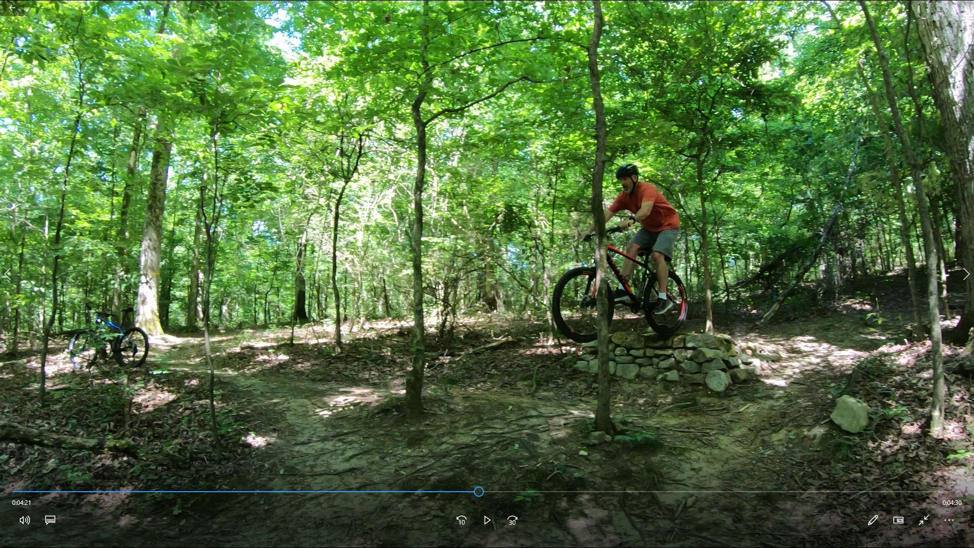 2017 Giant Talon 3: A mountain biker performing a trick on a stone feature in a lush green forest trail. The biker is in a red shirt and helmet, while another bike is parked nearby on the trail. Sunlight filters through the trees, creating a vibrant and lively outdoor scene.