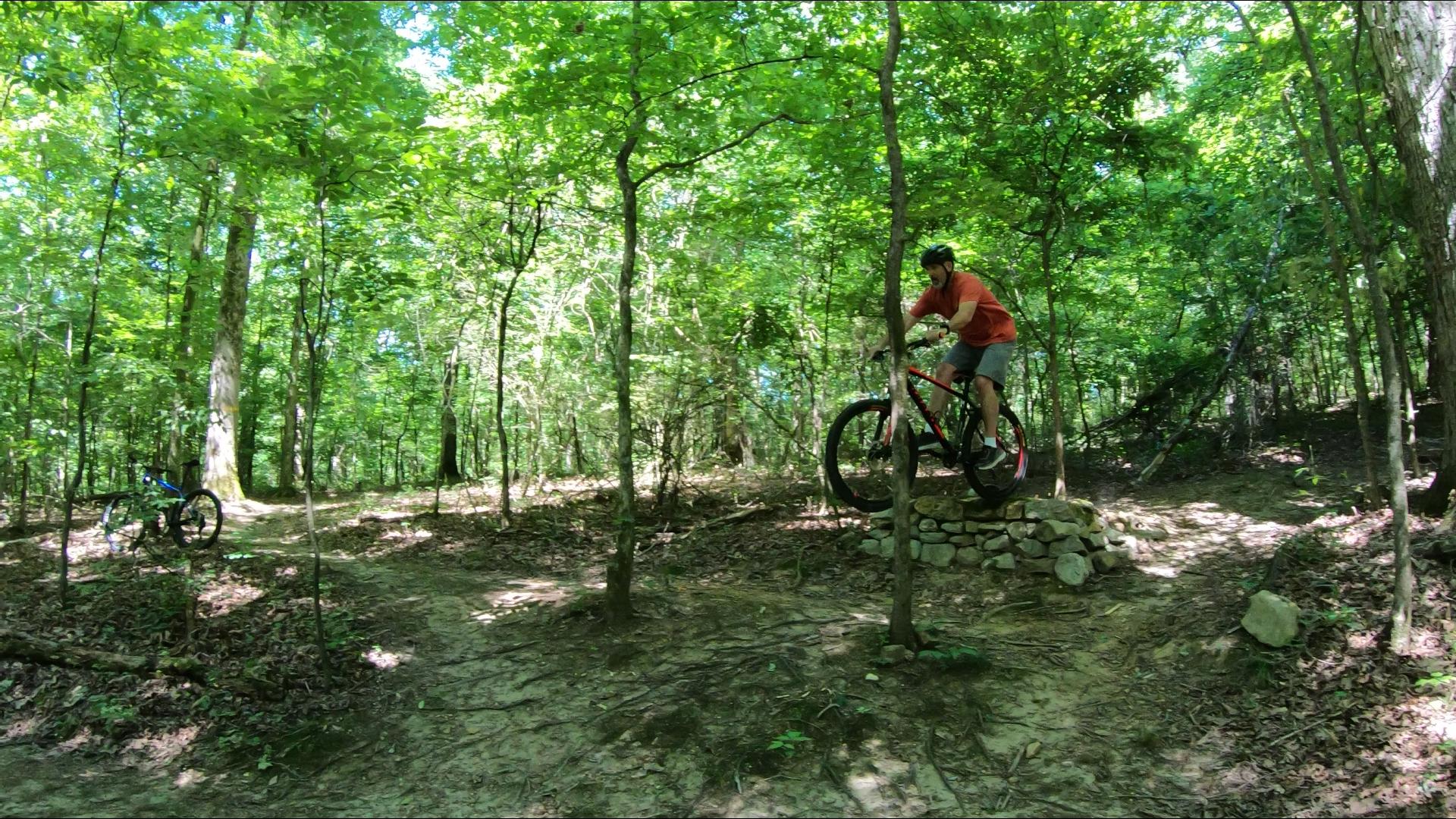 2017 Giant Talon 3: A mountain biker performing a tire lift on a stone feature in a lush green forest, surrounded by trees. In the background, a second bike is parked on the trail. The scene captures the thrill of outdoor biking and the beauty of nature.