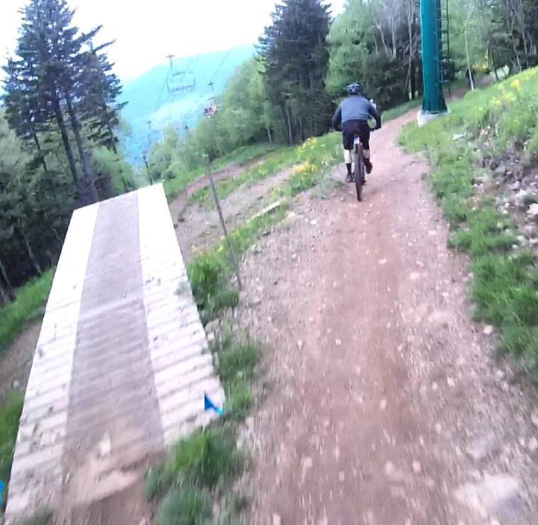 A mountain biker riding down a dirt trail near a wooden jump ramp, surrounded by trees and a scenic mountainous landscape. Snowshoe Bike Park mountain bike trail.