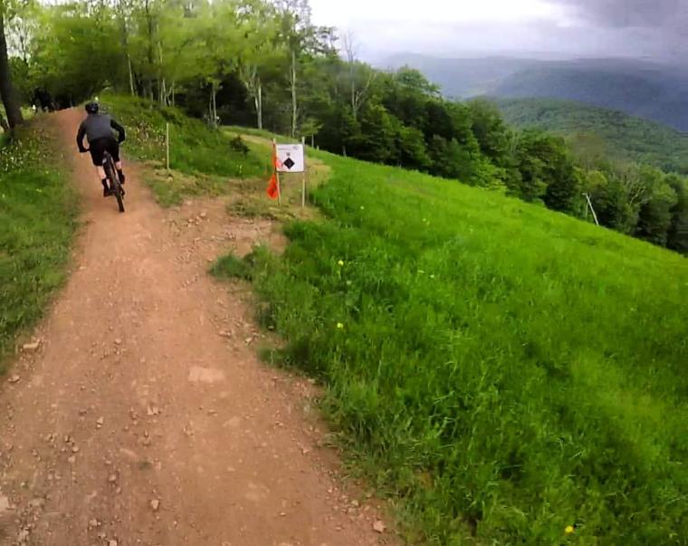 A person riding a mountain bike on a dirt path surrounded by lush green grass and trees. A warning sign with an orange flag is visible along the trail, indicating potential hazards ahead. In the background, rolling hills extend toward a cloudy sky. Snowshoe Bike Park mountain bike trail.