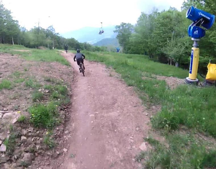 A mountain bike rider navigating a dirt trail on a grassy hillside, with a ski lift visible in the background. The scene is set in a green landscape with trees and mountains under a cloudy sky. Snowshoe Bike Park mountain bike trail.