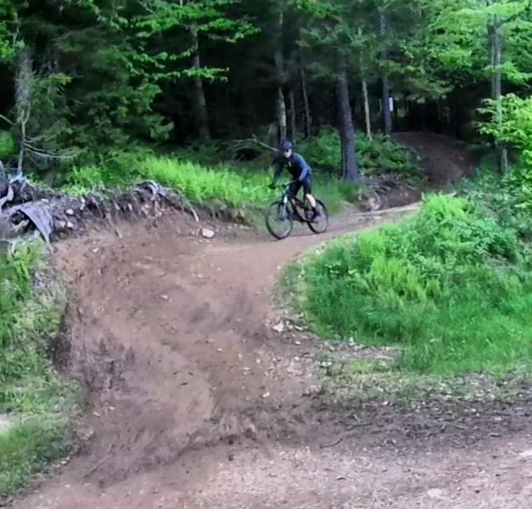 A mountain biker navigating a dirt trail in a wooded area, surrounded by lush greenery and trees. The trail curves to the right, showing a well-used path with some loose dirt and rocks. Snowshoe Bike Park mountain bike trail.