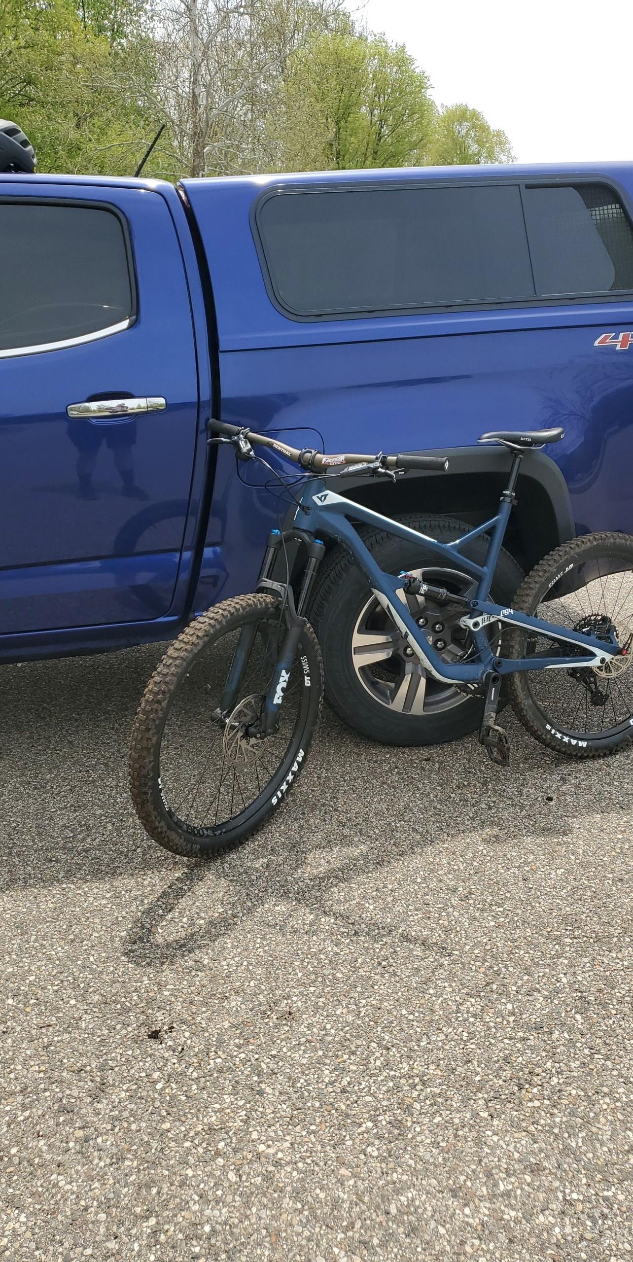 Yt industries Jeffsy: A blue mountain bike leaning against a blue pickup truck, parked on a gravel surface with trees in the background.