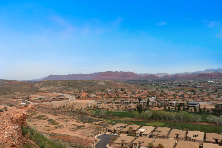 A panoramic view of residential homes set against a backdrop of rolling hills and mountains under a clear blue sky. The foreground features houses with beige rooftops, while the landscape extends into the distance, showcasing a mix of vegetation and arid land.