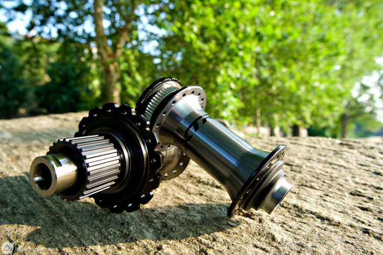 Close-up of a bicycle hub resting on a stone surface, surrounded by green trees. The hub has a shiny metallic finish with black accents and is positioned at an angle to showcase its details.