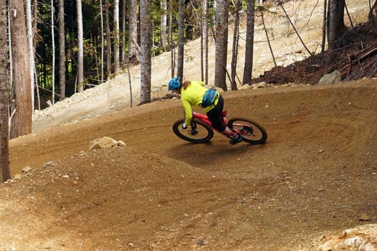 A mountain biker in a bright yellow jacket and blue helmet leans into a dirt turn on a forest trail, surrounded by tall trees and uneven terrain. The biker is navigating a winding path, showcasing an action-packed moment of a cycling adventure in nature.