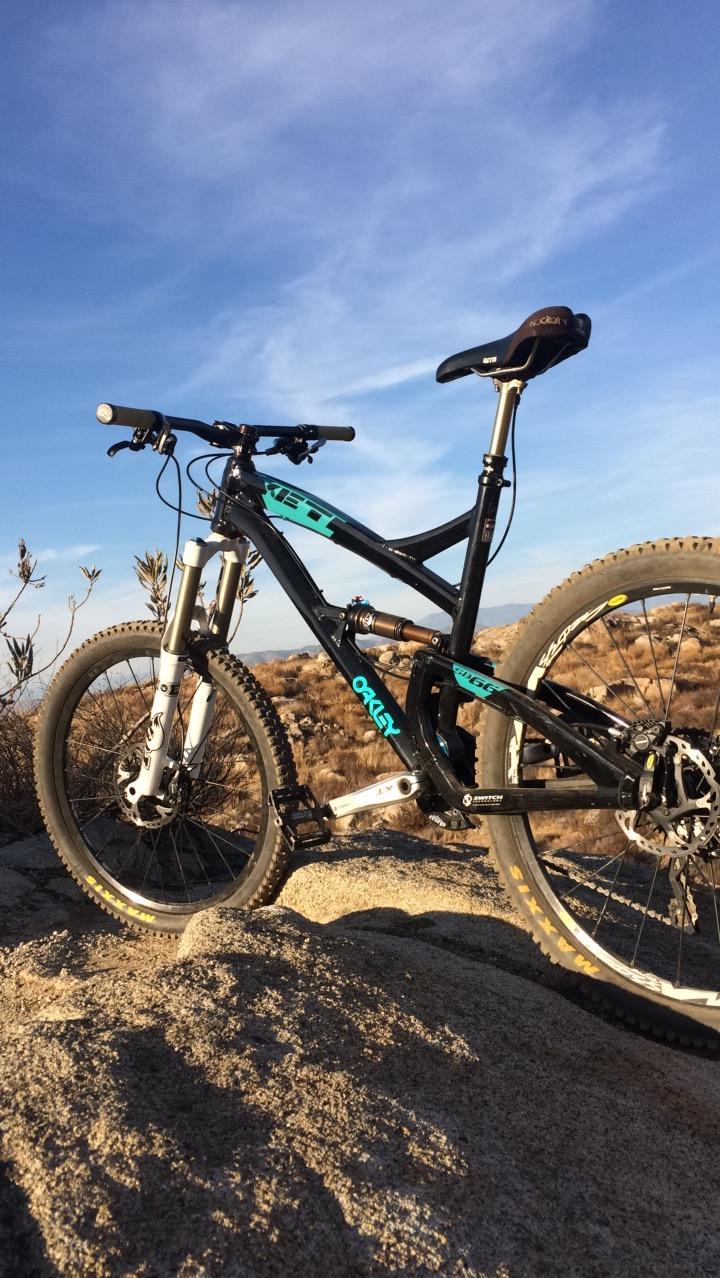 Yeti SB66: A black and turquoise mountain bike is positioned on rocky terrain under a clear blue sky. The bike features dual suspension, front and rear disc brakes, and rugged tires, indicating it's designed for off-road cycling. In the background, sparse shrubs and rolling hills create a natural outdoor setting.