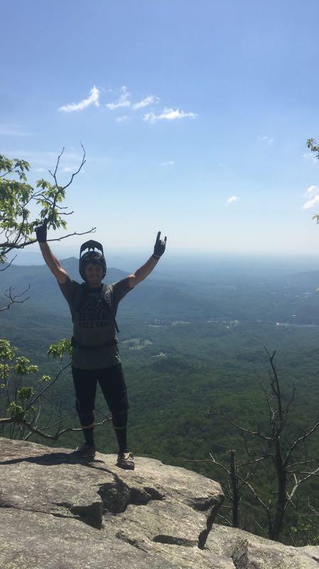 A hiker standing on a rocky outcrop with arms raised in celebration, overlooking a lush green valley and distant mountains under a clear blue sky. The individual is wearing a beanie and gloves, dressed in a casual athletic outfit. Weed Patch Mountain Trail mountain bike trail.