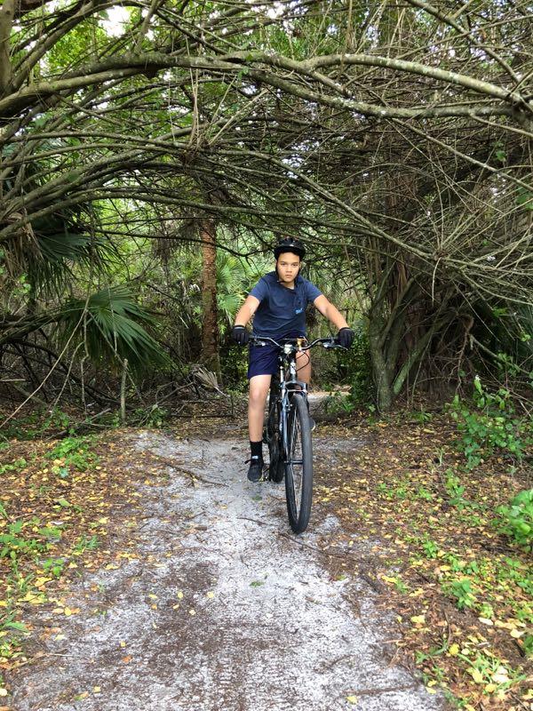 A child rides a bicycle along a dirt path surrounded by dense vegetation, including trees and shrubs, creating a natural archway above. The child is wearing a helmet and sport attire, focused on the ride as they navigate the trail. Okeeheelee Park / Pinehurst / Green Acres Freedom Park mountain bike trail.