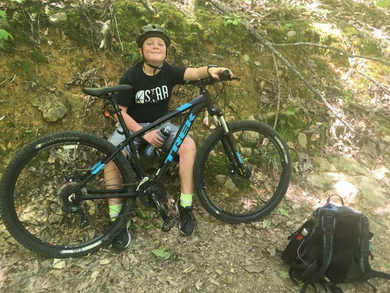 A young person wearing a helmet sits next to a black mountain bike on a dirt trail surrounded by greenery. They are smiling and holding a water bottle, with a backpack resting on the ground nearby. Coldwater Mountain mountain bike trail.