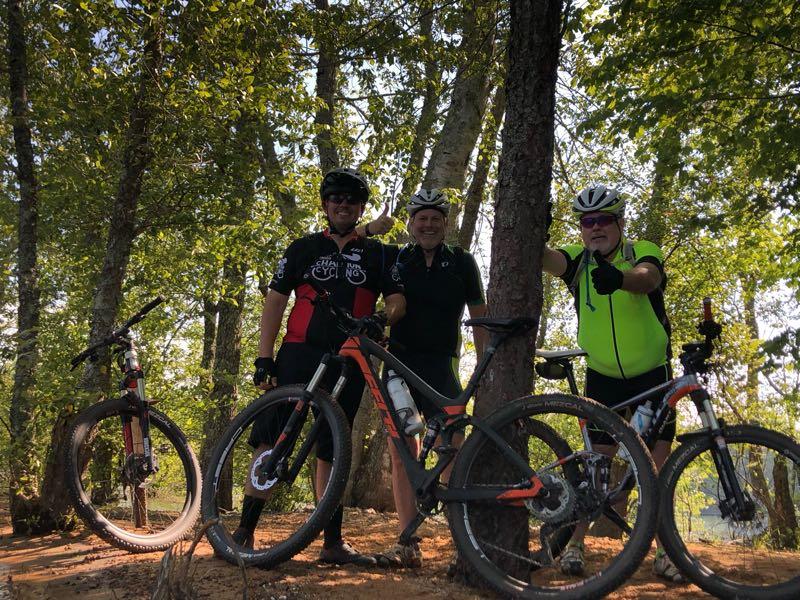 Three cyclists posing together next to their mountain bikes in a wooded area. They are smiling and wearing cycling gear, with trees providing a lush green backdrop. Jack Rabbit Trails mountain bike trail.