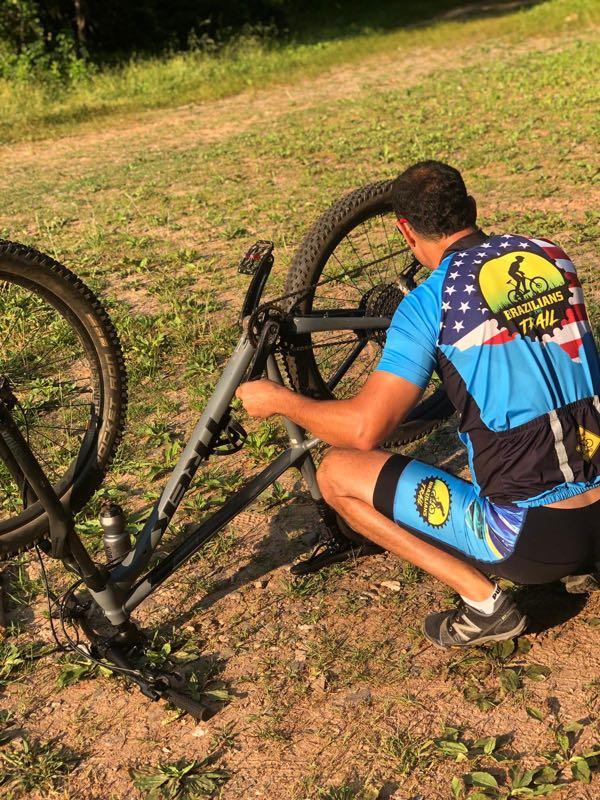 A person wearing a colorful cycling jersey and shorts is kneeling beside a mountain bike on a dirt path, adjusting the bike's rear wheel. The background features a grassy area and trees, indicating an outdoor setting ideal for biking. Bull / Jake Mountain mountain bike trail.