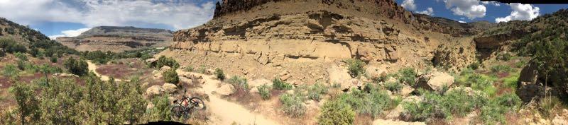 A panoramic view of a rugged landscape featuring towering cliffs, a winding dirt trail, and vibrant green vegetation. The sky is dotted with clouds, and the terrain showcases a mix of rocks and shrubs, highlighting the natural beauty of the area. A bicycle is partially visible on the trail, suggesting an outdoor adventure scene. Palisade Rim mountain bike trail.