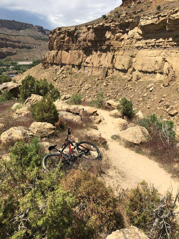 A mountain bike rests on a dirt trail surrounded by rocky terrain and sparse vegetation, with steep cliffs and hills in the background under a partly cloudy sky. Palisade Rim mountain bike trail.