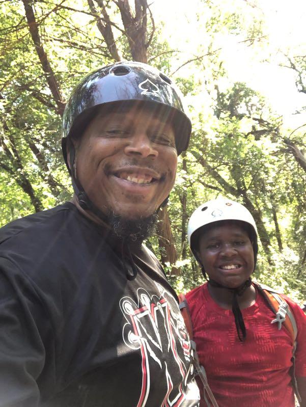 A smiling adult and child wearing helmets, standing together outdoors surrounded by trees. The sun is shining through the foliage, creating a warm and cheerful atmosphere. The adult is wearing a black shirt with a graphic design, while the child is dressed in a bright red shirt. Cedar Hill State Park At Joe Pool Lake mountain bike trail.