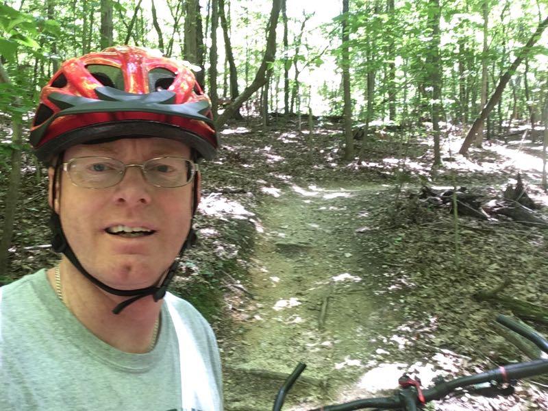 A person in a red bike helmet, taking a selfie on a dirt mountain biking trail surrounded by vibrant green trees. The path in the background suggests an outdoor recreational area. Country Park mountain bike trail.