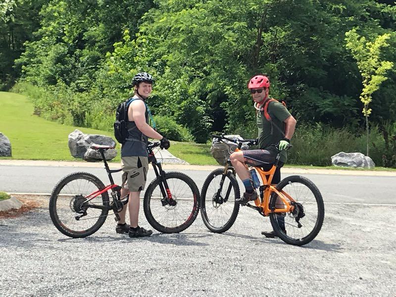 Two young men stand on a gravel path next to their mountain bikes. One is wearing a black helmet, a sleeveless blue shirt, and tan shorts, while the other is wearing a red helmet, a green shirt, and gray shorts. They both smile and face the camera against a backdrop of green trees and rocks. Rappahanock River Trail mountain bike trail.