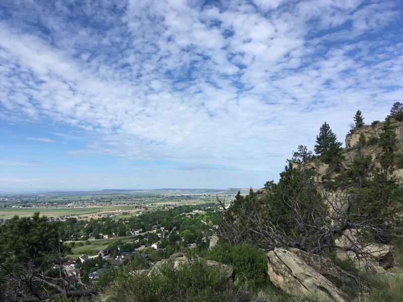 A panoramic view of a green valley surrounded by rocky hills under a partly cloudy sky. In the foreground, there are shrubs and trees, while the valley below features a mix of residential areas and open fields. The horizon is visible in the distance, creating a serene natural landscape. Zimmerman Trail mountain bike trail.