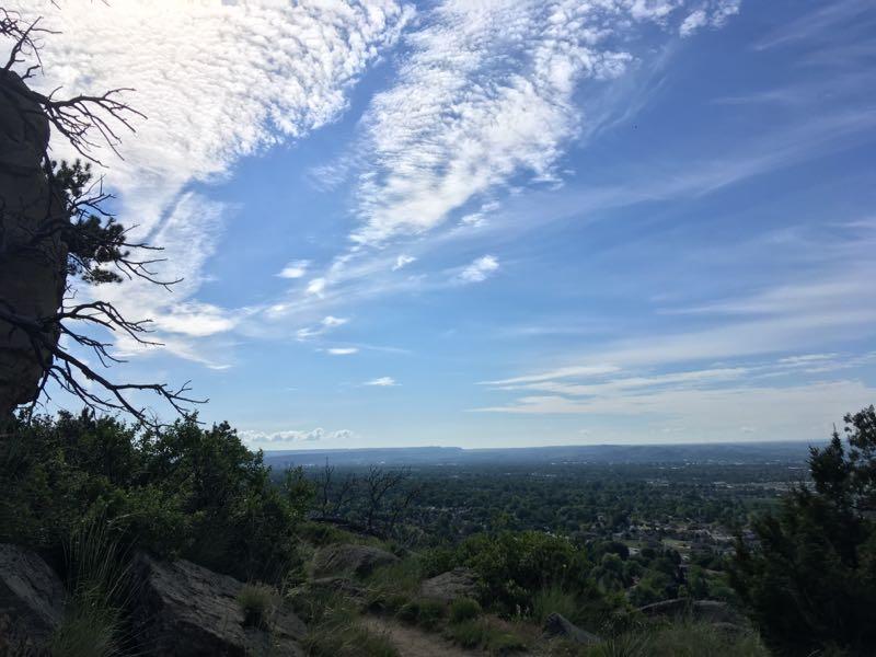 A panoramic view of a landscape featuring a mix of rocky outcrops, green vegetation, and a blue sky filled with clouds. The scene captures the natural beauty of the area, highlighting the horizon with distant hills in the background. Zimmerman Trail mountain bike trail.