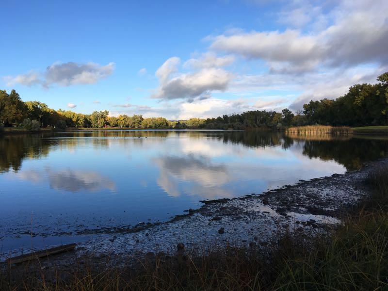 A tranquil lake reflecting the blue sky and clouds, surrounded by trees. The shoreline features patches of grass and mud, with a few small logs visible along the water's edge. The scene conveys a calm, peaceful natural setting. River Front Park mountain bike trail.